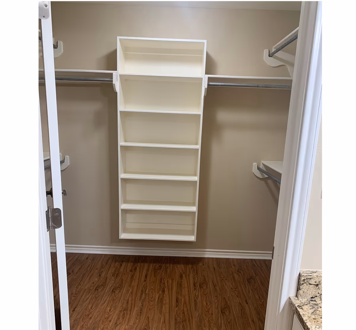 Empty walk-in closet with wooden floor, beige walls, white shelving unit with multiple compartments in the center, and metal hanging rods on both sides.