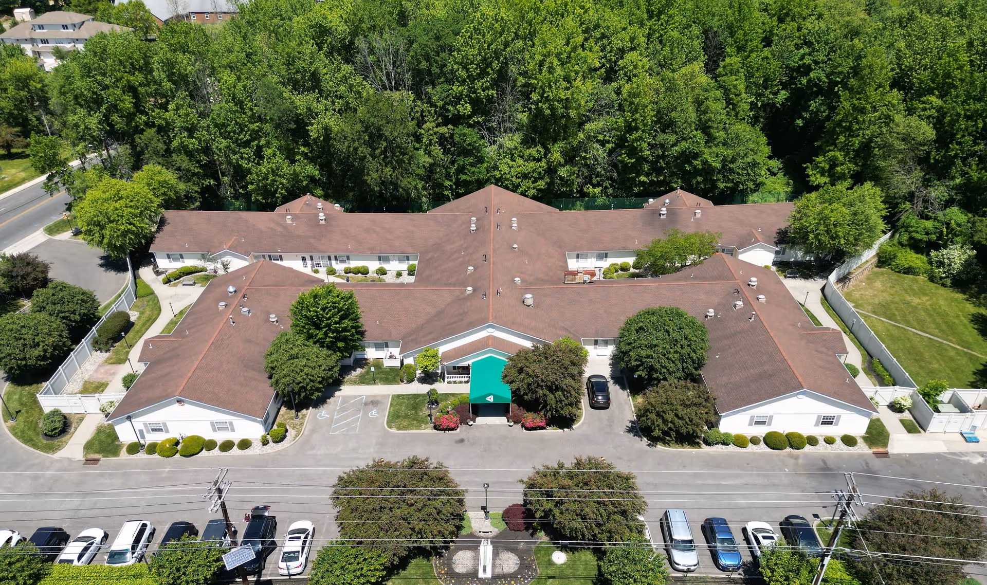 Aerial view of Arden Courts - ProMedica Memory Care Community in Cherry Hill, showing a large single-story building with a brown roof surrounded by trees and greenery. There is a green canopy over the main entrance, parking spaces with several cars in front, and a road running parallel to the building.