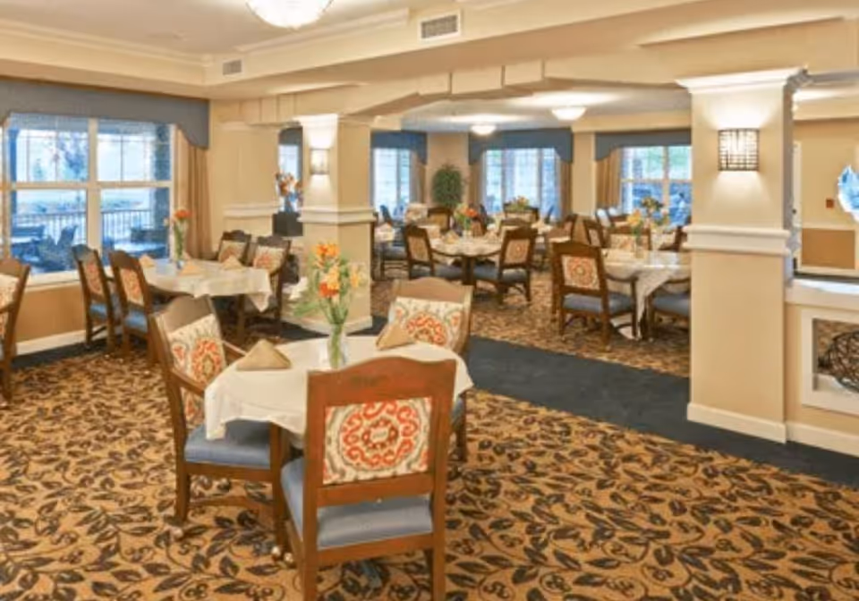 Bright carpeted dining room with multiple round tables and chairs set with tablecloths and floral centerpieces.