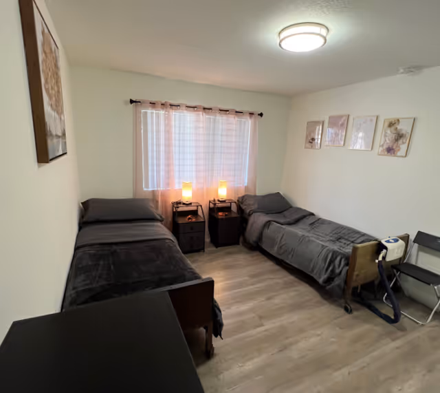 A small bedroom with two single beds placed parallel to each other against opposite walls. Each bed has dark gray bedding and a small bedside table with a lamp. A window with sheer curtains is centered between the beds. The room has light-colored walls and wood flooring. There are framed pictures on the wall above one bed and a chair with a medical device next to the other bed.