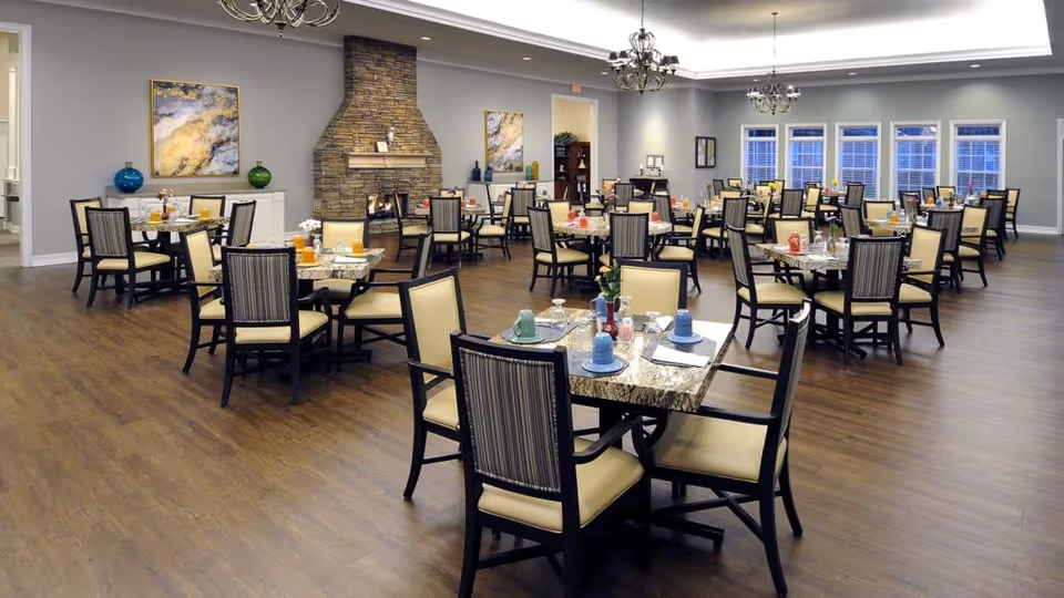 A spacious dining room with multiple tables and chairs arranged neatly. Each table is set with plates, cups, and utensils. The room features a stone fireplace in the background, two abstract paintings on the walls, and large windows letting in natural light. The floor is wooden, and chandeliers hang from the ceiling providing warm lighting.