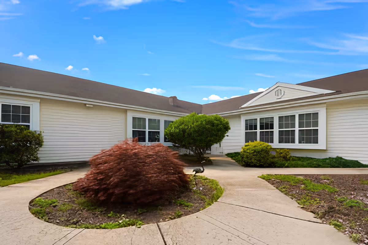 Single-story white building with a central walkway, landscaped shrubs, and large windows under a blue sky.