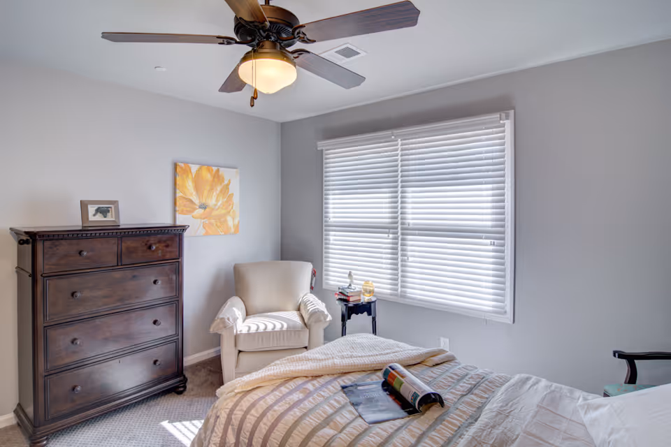 Sunlit bedroom with a bed, armchair, dark wooden dresser, ceiling fan, and window blinds.