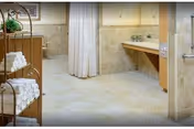 A clean and spacious bathroom area with beige tiled floors and walls. There is a wooden shelving unit on the left holding neatly folded white towels. A shower area with a white curtain is visible in the background, and a long countertop with a sink and a mirror is on the right side. The space is well-lit and has a calm, neutral color scheme.