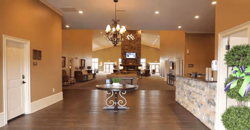 Interior view of a senior living facility lobby with a round table in the center holding decorative items, a stone reception desk on the right, and a large stone fireplace with a mounted TV in the background. The space is warmly lit with chandeliers and recessed lighting, featuring comfortable seating areas and beige walls.