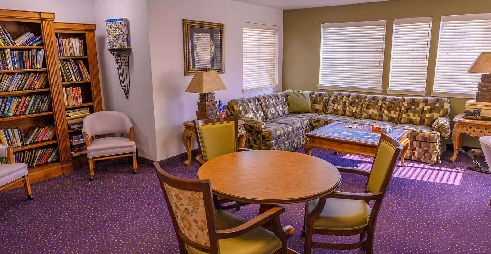 Community lounge with a round table and chairs, bookshelf, patterned sectional sofa and coffee table in front of windows.