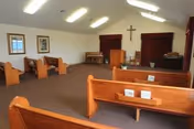 Interior view of a small chapel or worship room with wooden pews arranged facing a podium and piano. The room has a high ceiling with fluorescent lights, a cross mounted on the wall behind the podium, and framed pictures on the side walls.