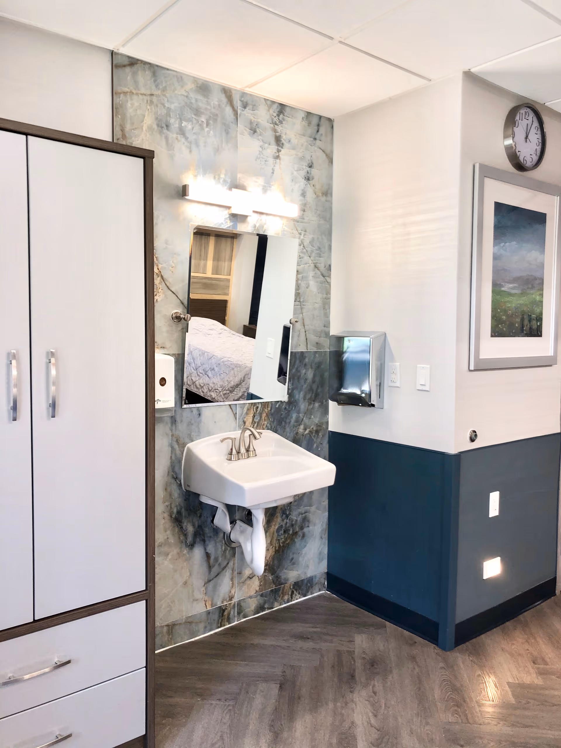 A small sink mounted on a marble-patterned wall with a mirror above it reflecting part of a bed. To the left is a white cabinet with drawers and silver handles. On the right wall, there is a metal paper towel dispenser, a clock, and a framed landscape painting. The floor has a wood-like pattern.
