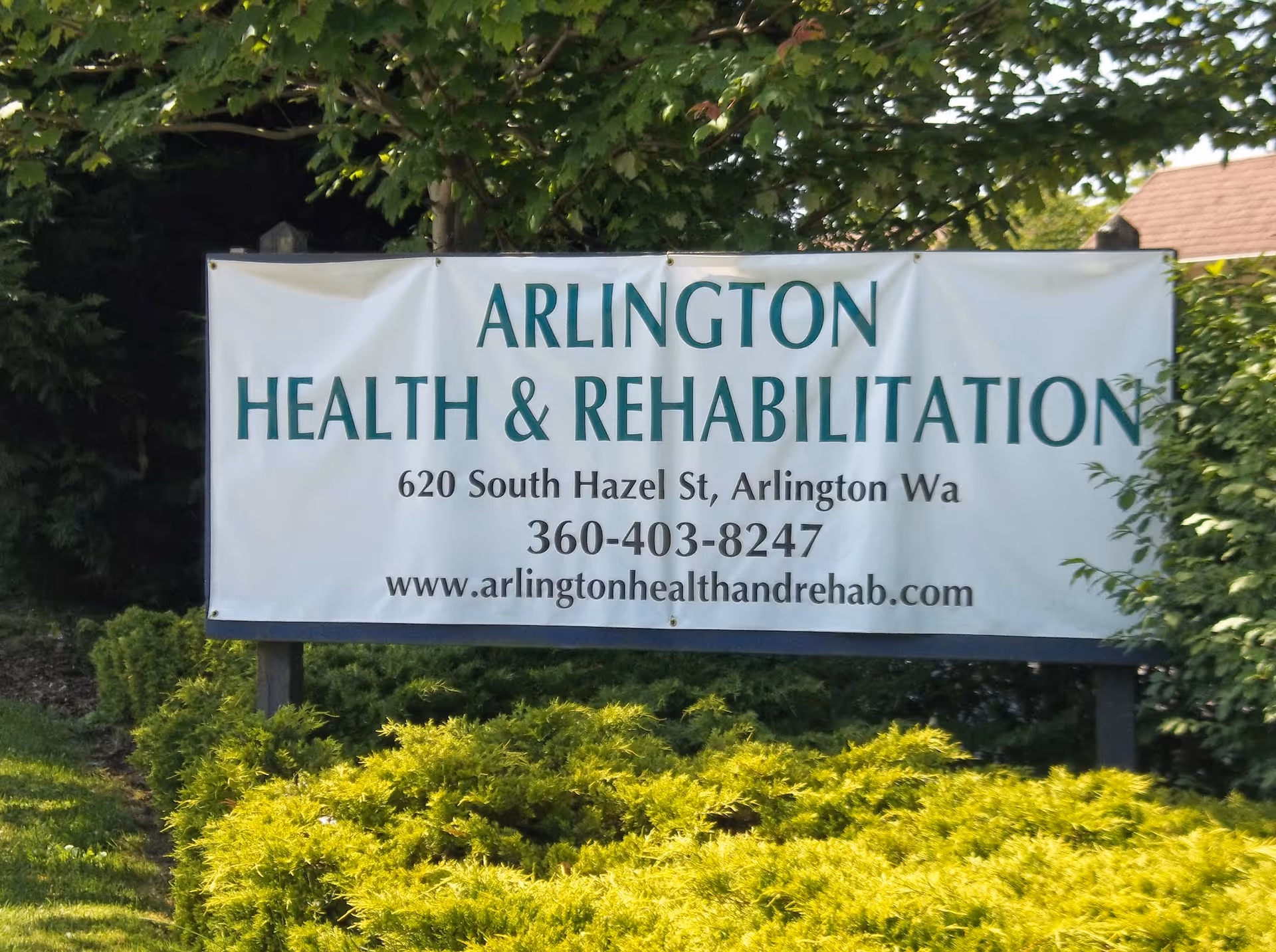 Outdoor sign for Arlington Health & Rehabilitation located at 620 South Hazel St, Arlington, Washington, with contact number 360-403-8247 and website www.arlingtonhealthandrehab.com, surrounded by green bushes and trees.