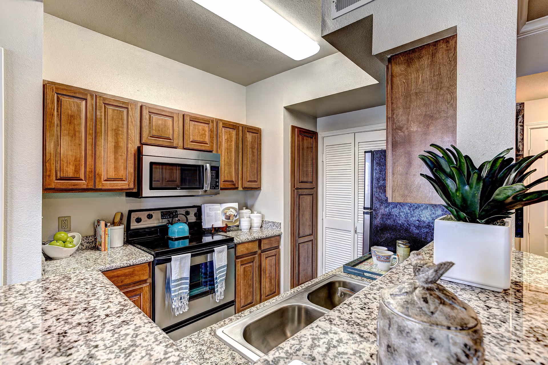 Modern kitchen with granite countertops, wooden cabinets, stainless steel microwave and stove, a double sink, and decorative items including a bowl of green apples, a teal kettle, and a potted plant.