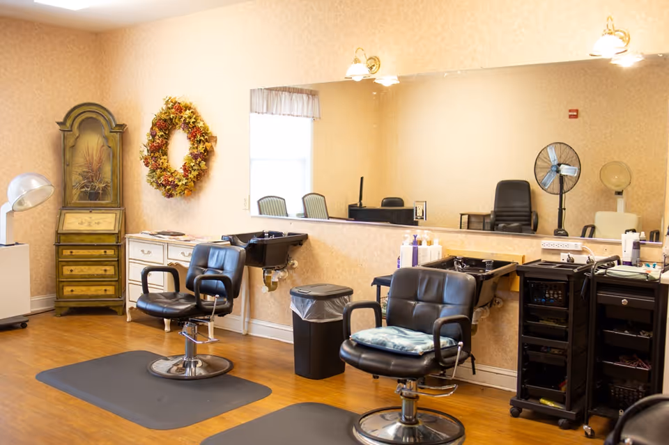 A hair salon room with styling chairs, wash basins, a large mirror, and hair-drying equipment.