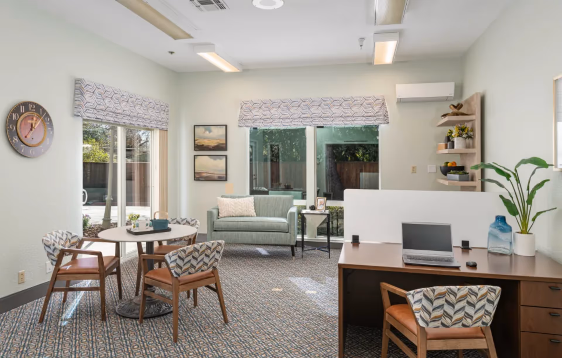 A bright and cozy senior living common area with a small round table surrounded by four patterned chairs, a green loveseat with a white pillow, a side table with a framed photo, and a wooden desk with a laptop, mouse, and a plant. Large windows with patterned valances let in natural light, and the room has a patterned carpet and light-colored walls.