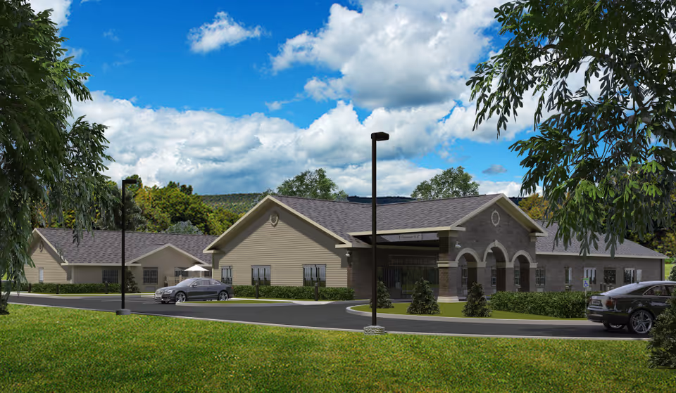 Front exterior view of a one-story senior living facility with a driveway, parked cars, and landscaped lawn under a blue sky.