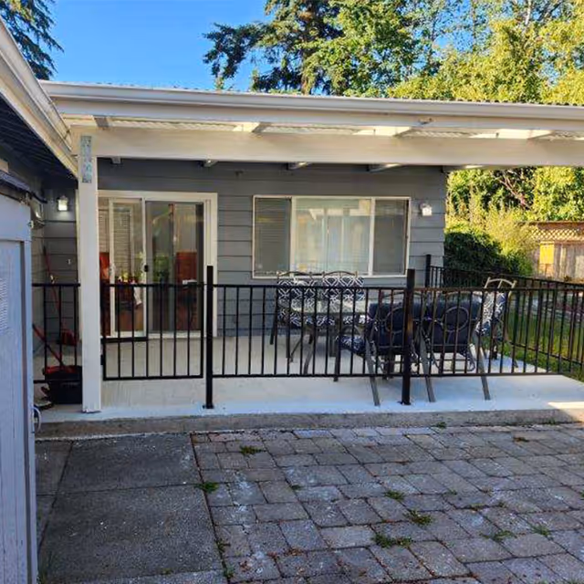 Covered backyard patio with a black metal railing and an outdoor table and chairs in front of a gray single-story house.