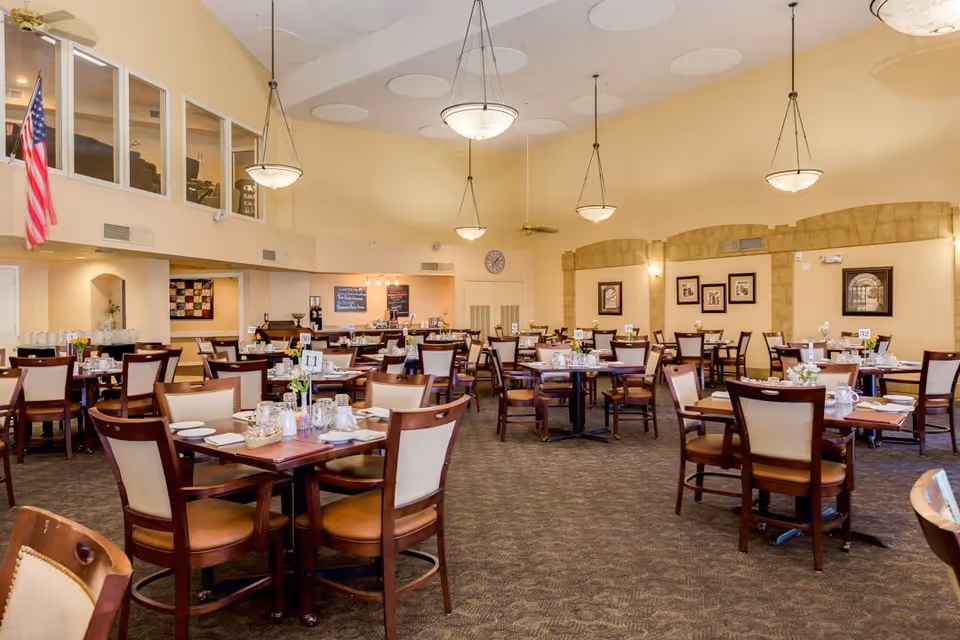 A spacious dining room with multiple wooden tables and chairs arranged neatly. Each table is set with plates, glasses, and utensils, and small flower arrangements are placed on the tables. The room has high ceilings with hanging light fixtures and beige walls adorned with framed pictures. An American flag is visible on the left side near a windowed area above a counter.