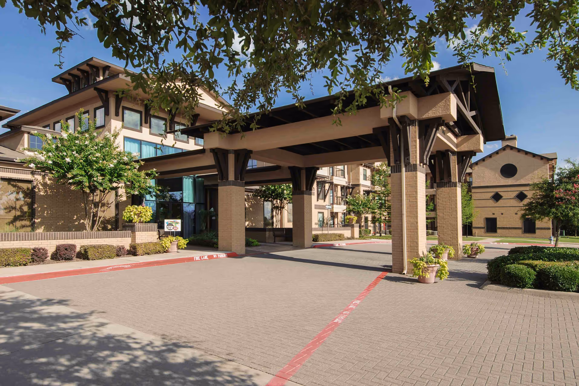 Exterior view of Parkview in Allen senior living facility showing a covered entrance with large pillars, surrounded by trees and shrubs under a clear blue sky.