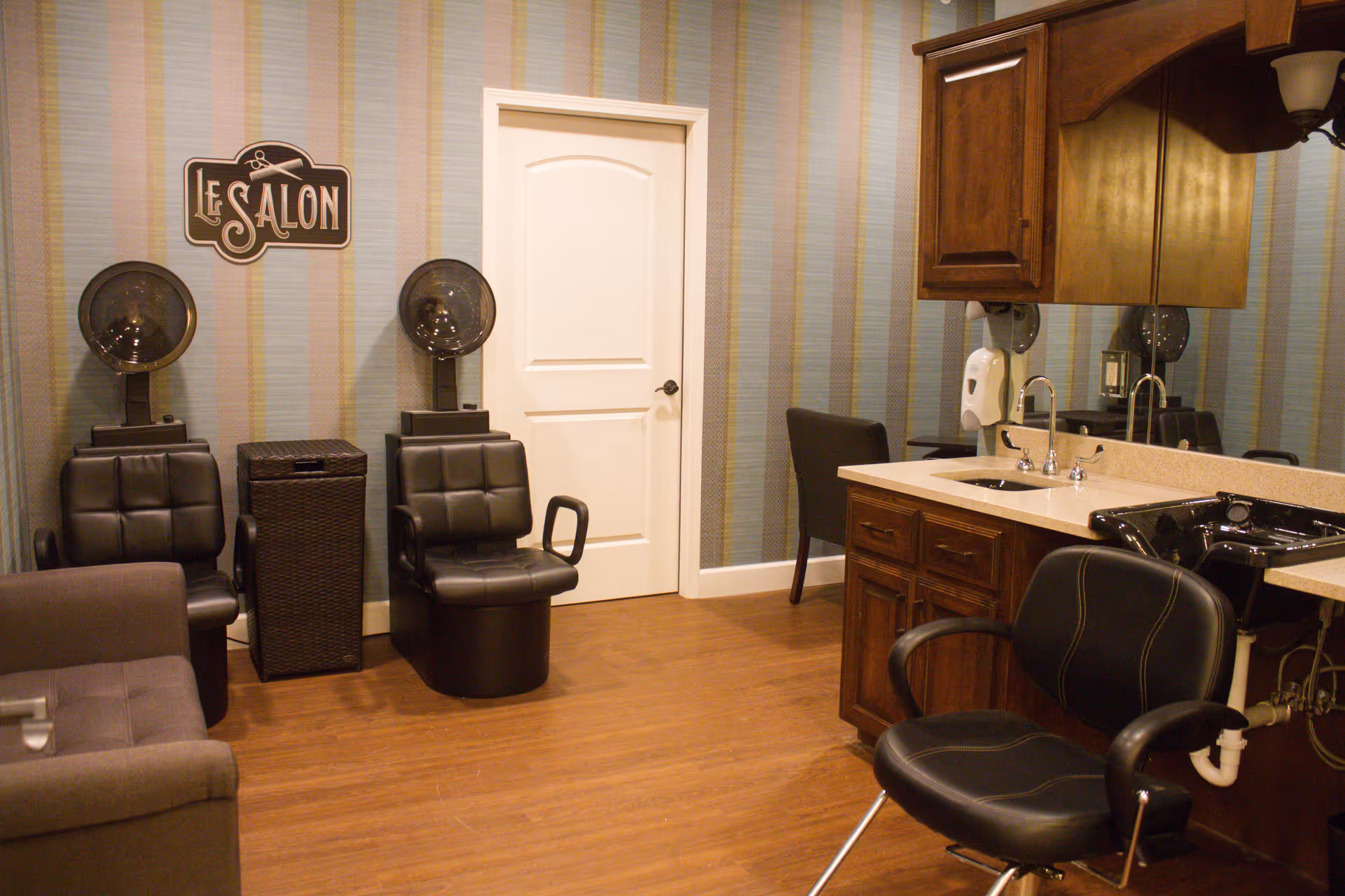 Interior of a salon room with two black salon chairs under hair dryers, a wicker laundry basket between them, a white door in the background, a brown cushioned chair in the foreground, and a countertop with a sink and a black salon chair in front of a large mirror.