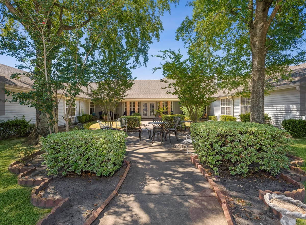 Outdoor courtyard area at Rosefield Court featuring a paved walkway lined with trimmed bushes and trees. Several metal chairs and tables are arranged in the center for seating. The building with white siding and multiple windows surrounds the courtyard under a clear blue sky.
