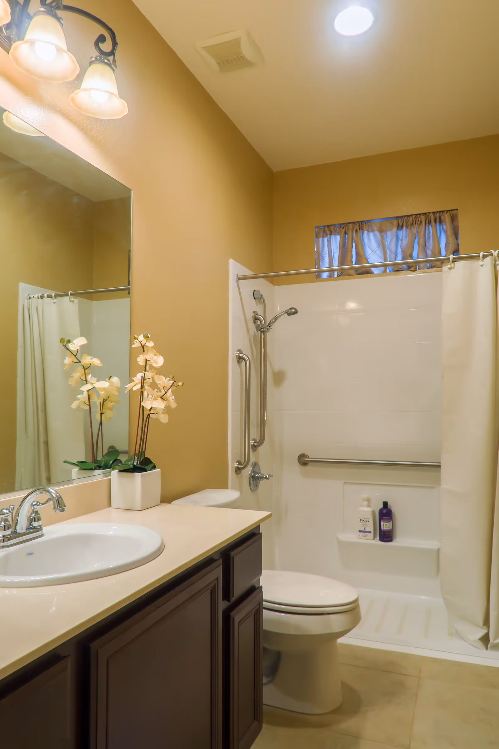 Well-lit bathroom with a sink and vanity, toilet, and a shower/tub with grab bars and curtain.