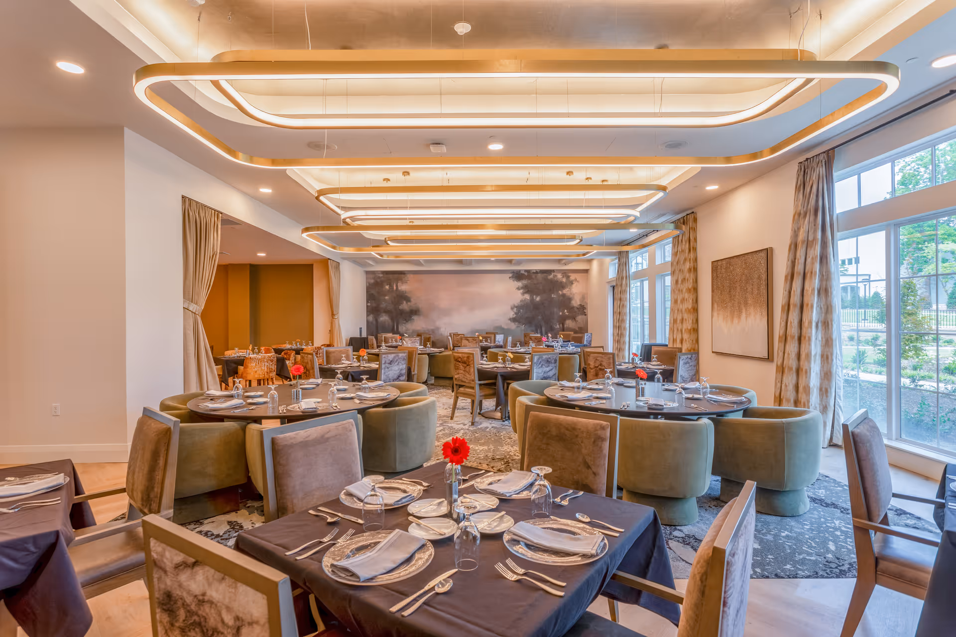 A spacious, well-lit dining room with multiple round and square tables set with plates, napkins, glasses, and silverware. The room features modern ceiling light fixtures, large windows with curtains allowing natural light, and a mural on the far wall depicting trees and a misty landscape.