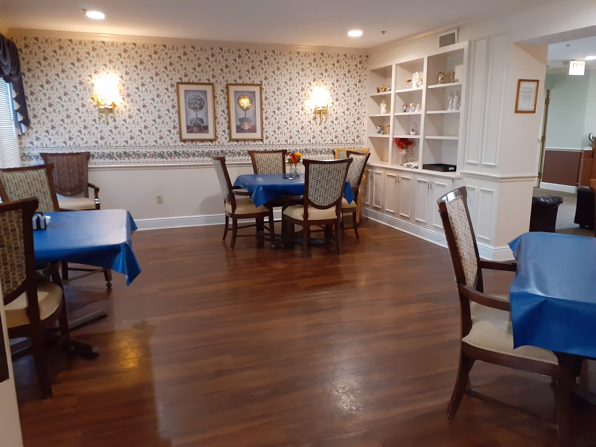 A dining room with wooden floors and several tables covered with blue tablecloths. Each table is surrounded by wooden chairs with cushioned seats and patterned backs. The walls have floral wallpaper with two framed pictures and wall sconces providing light. There is a built-in white cabinet with shelves displaying decorative items on the right side of the room.