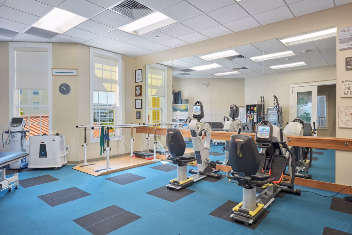 A bright exercise room with large windows, blue and black checkered carpet, and various fitness equipment including stationary recumbent bikes, parallel bars for walking support, and other rehabilitation machines. The room has a large mirror on one wall and motivational wall art.