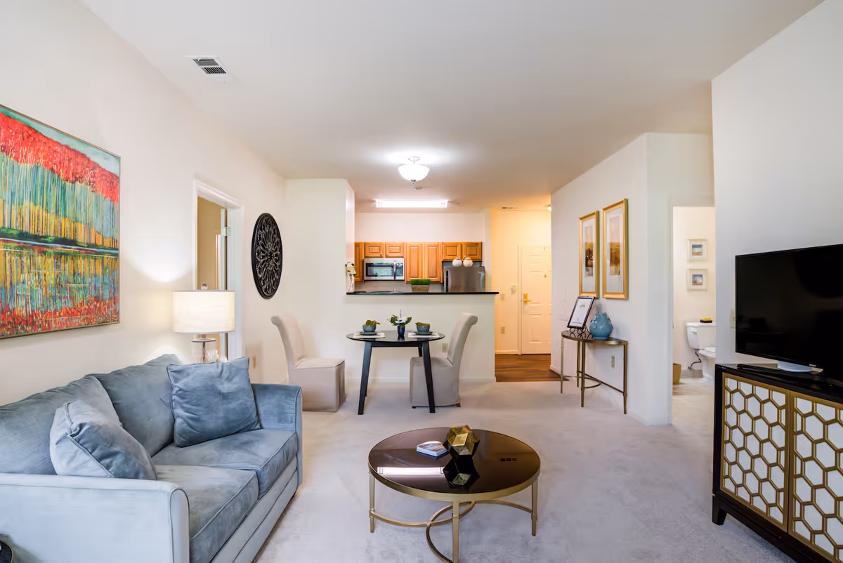 A bright and clean living room area with a light blue sofa, a round coffee table, and a flat-screen TV on a decorative stand. There is a small dining table with two chairs near the kitchen, which has wooden cabinets and stainless steel appliances. The walls are decorated with colorful artwork and framed pictures. A doorway leads to a bathroom with a visible toilet.