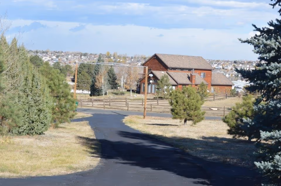 A paved driveway curves through a grassy yard with trees toward a two-story rustic wooden house and fence under a partly cloudy sky.