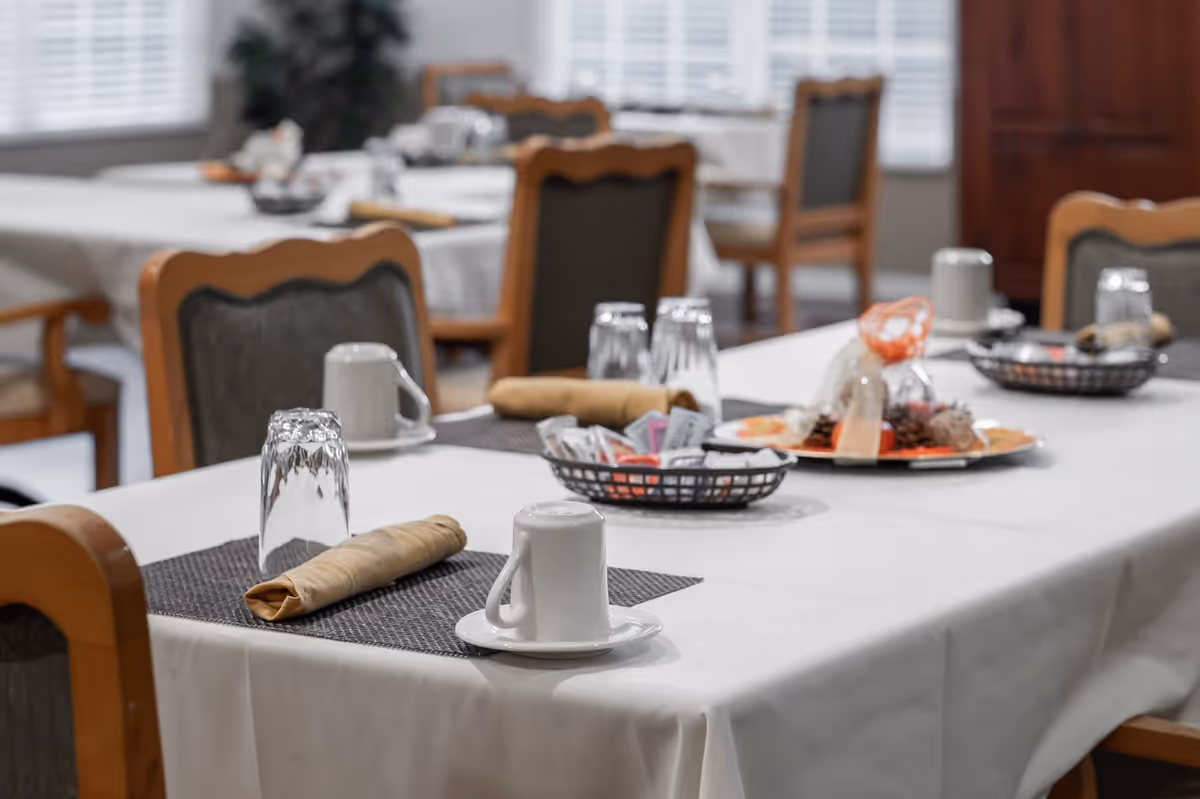 A dining table set with white tablecloths, beige napkins, upside-down glasses, white coffee cups on saucers, and baskets containing condiments and snacks in a well-lit room with wooden chairs.