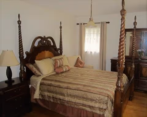 Traditional bedroom with an ornate four-poster wooden bed, bedside tables, lamp, dresser and window with curtains.