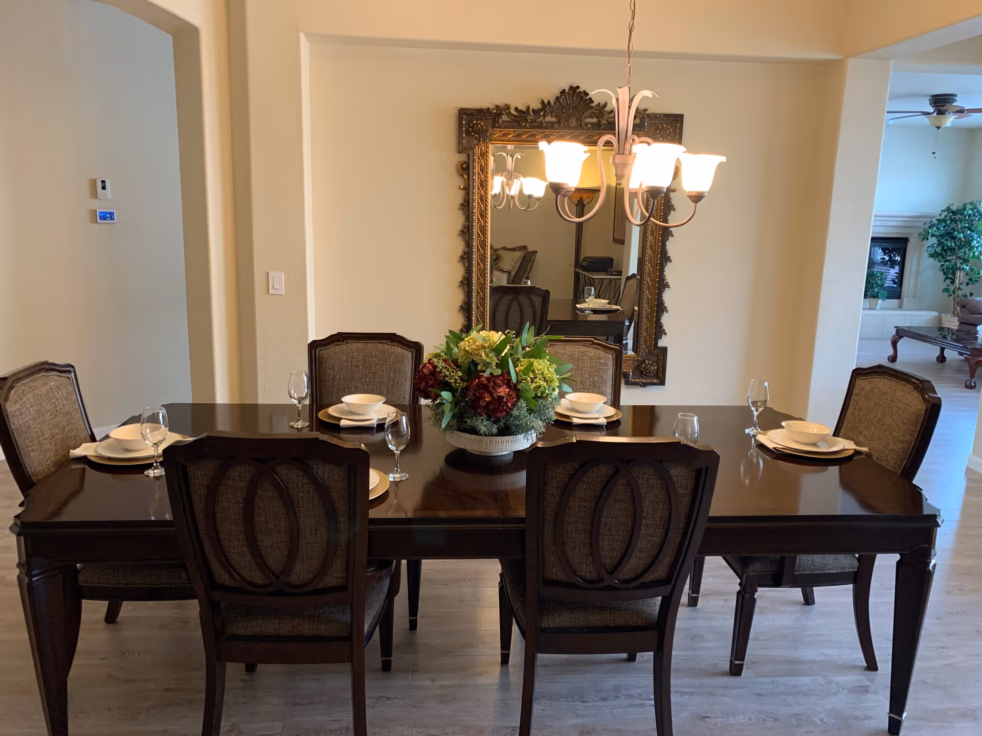 A dining room with a dark wooden table set for six people with plates, bowls, and wine glasses. A floral centerpiece is placed in the middle of the table. There is a large ornate mirror on the wall behind the table and a chandelier with six lights hanging above. The room has light-colored walls and flooring, and a living room area is visible in the background.