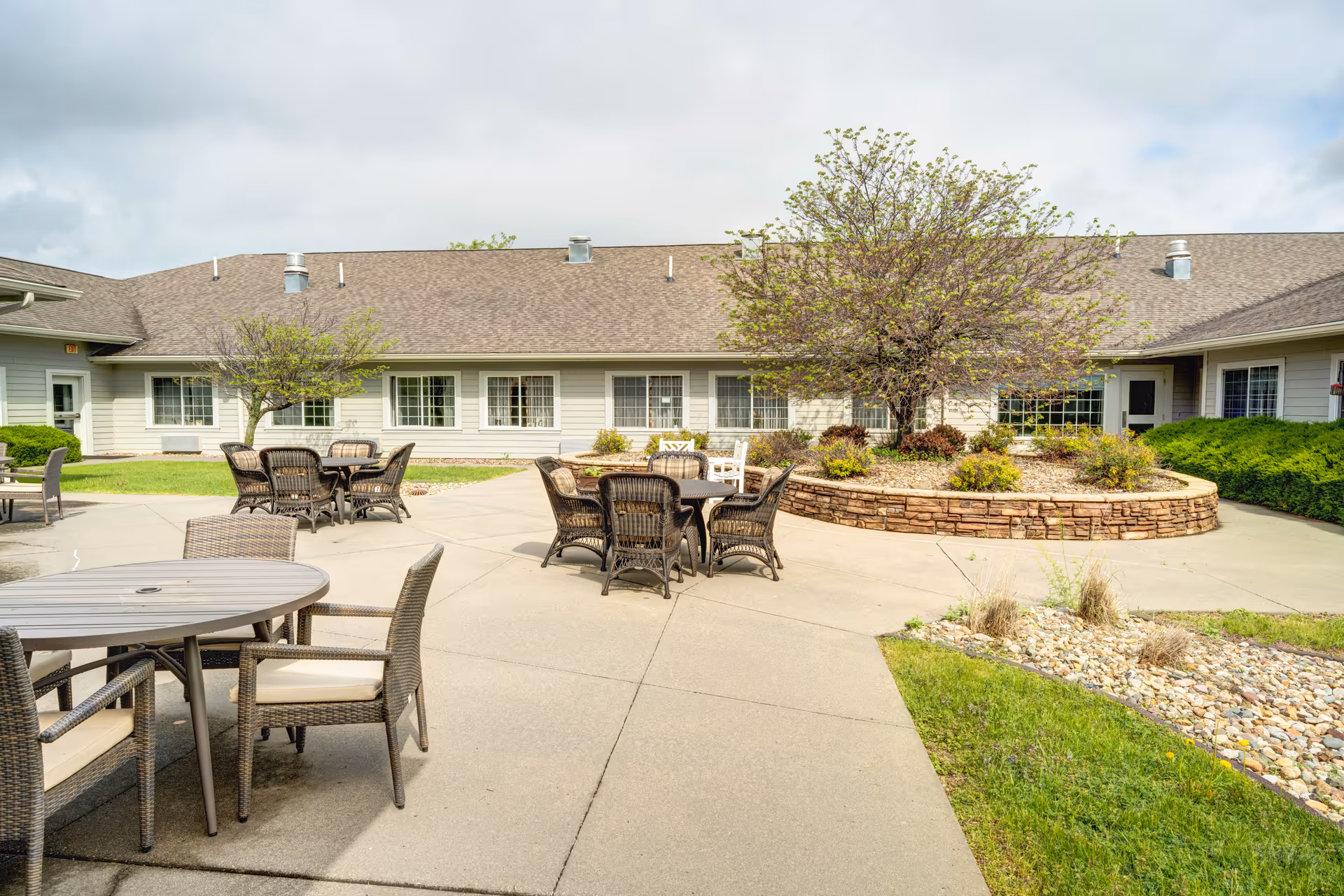 Outdoor patio area at a senior living facility with multiple round tables and wicker chairs arranged on a concrete surface. There is a raised circular garden bed with a tree and shrubs in the center, surrounded by a building with multiple windows and a sloped roof under a partly cloudy sky.