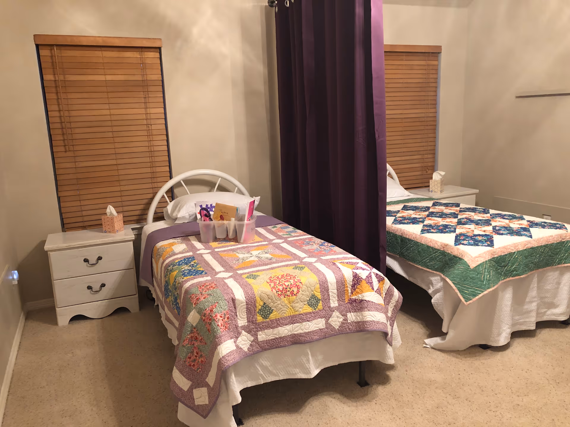 A bedroom with two single beds separated by a purple privacy curtain. Each bed has a colorful patchwork quilt, a white pillow, and a white nightstand with a box of tissues. The room has beige walls and carpet, and two windows with closed wooden blinds.
