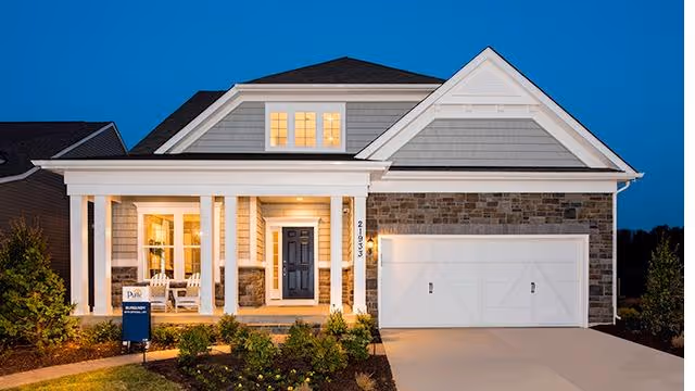 Exterior view of a single-story house at dusk with a well-lit front porch, white columns, a dark front door, and a two-car garage. The house has a combination of stone and siding on the facade, with a manicured lawn and shrubs in front.
