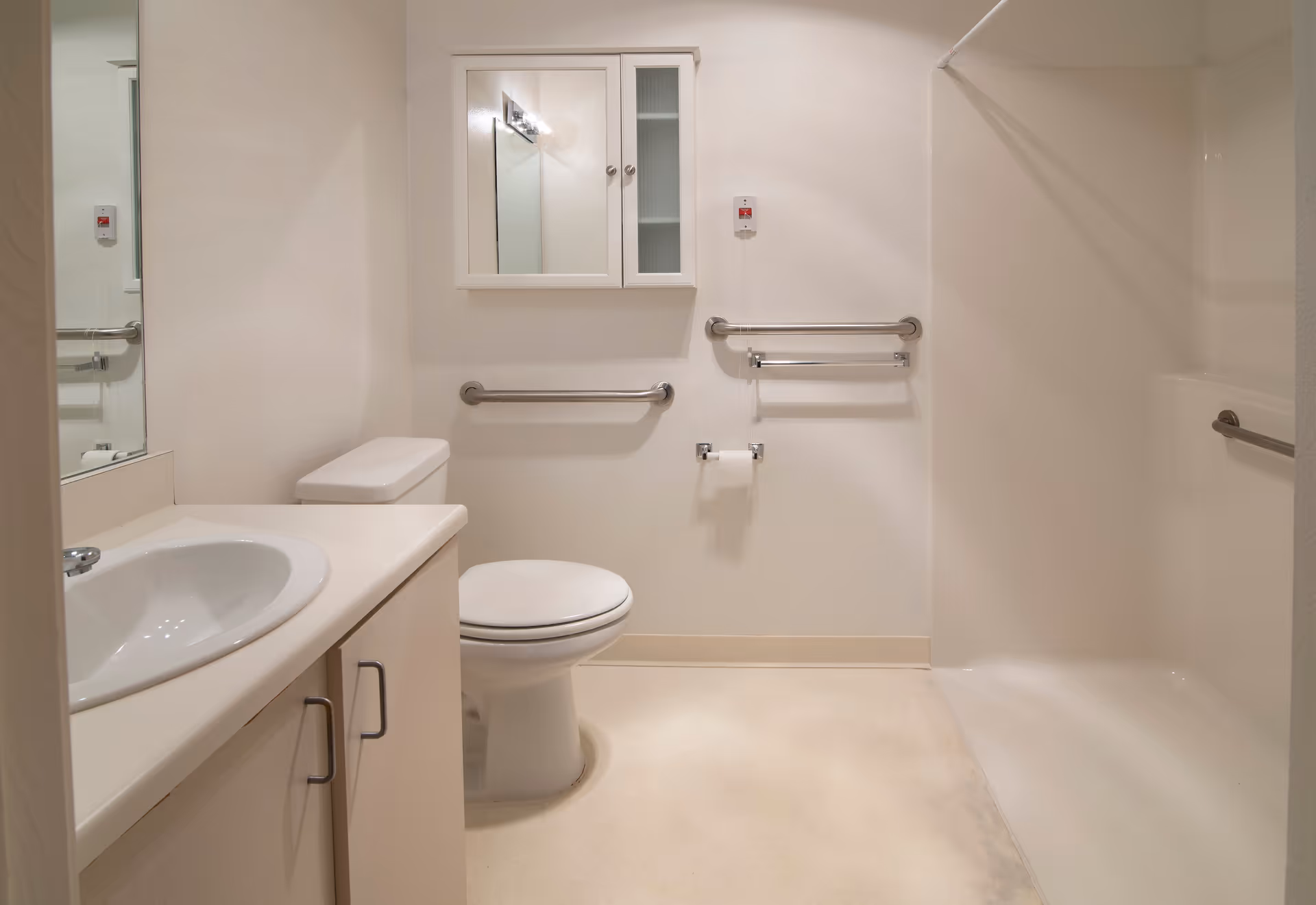 A clean, accessible bathroom with a white sink and countertop on the left, a toilet in the center, and a walk-in shower on the right. The walls and floor are light-colored, and there are multiple stainless steel grab bars installed near the toilet and inside the shower. A mirrored medicine cabinet is mounted on the wall above the toilet.