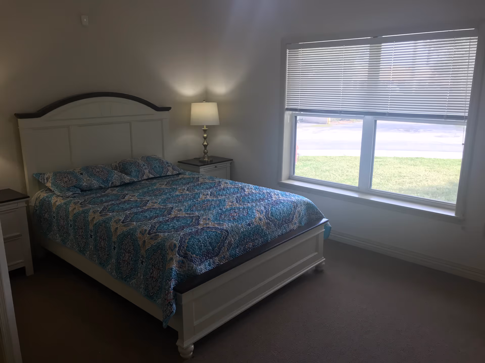 A bedroom with a white wooden bed frame and a blue patterned bedspread. There are two matching pillows on the bed. On each side of the bed are white nightstands, one with a lamp turned on. A large window with closed blinds lets in natural light, showing a grassy area outside.