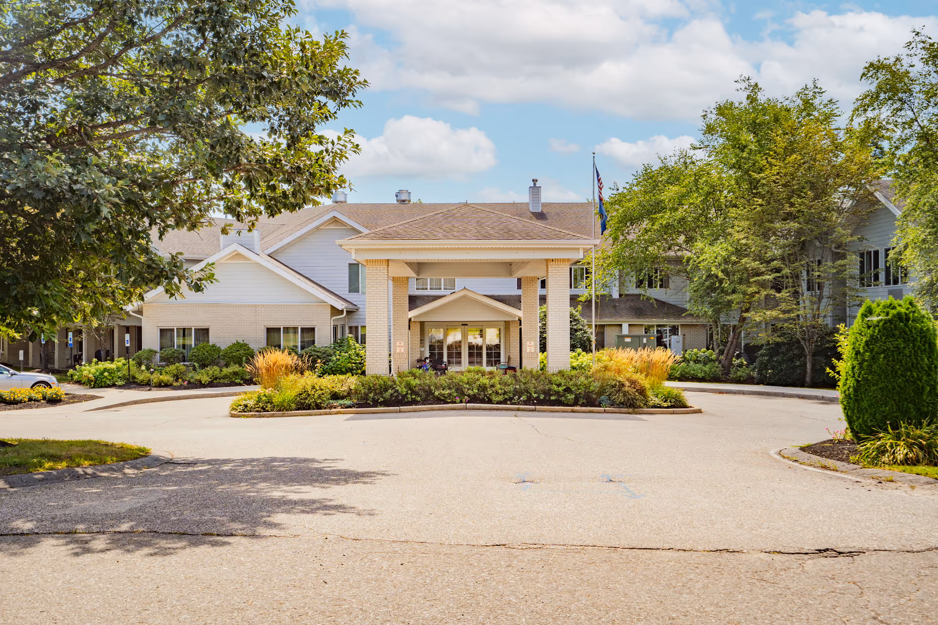Front exterior view of Birchwoods at Canco Senior Living facility showing a covered entrance with pillars, surrounded by landscaped greenery and trees under a partly cloudy sky.