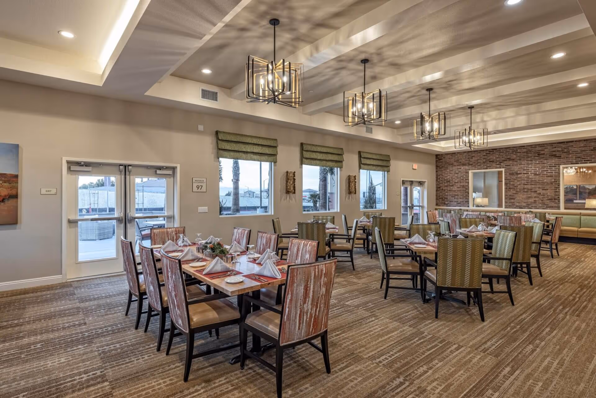 A spacious dining room in a senior living facility with multiple tables set with white napkins, plates, and cutlery. The room features patterned chairs, large windows with green roman shades, modern chandeliers, and a brick accent wall with a large mirror. The carpet has a neutral pattern, and there are exit doors visible on the left and right sides.