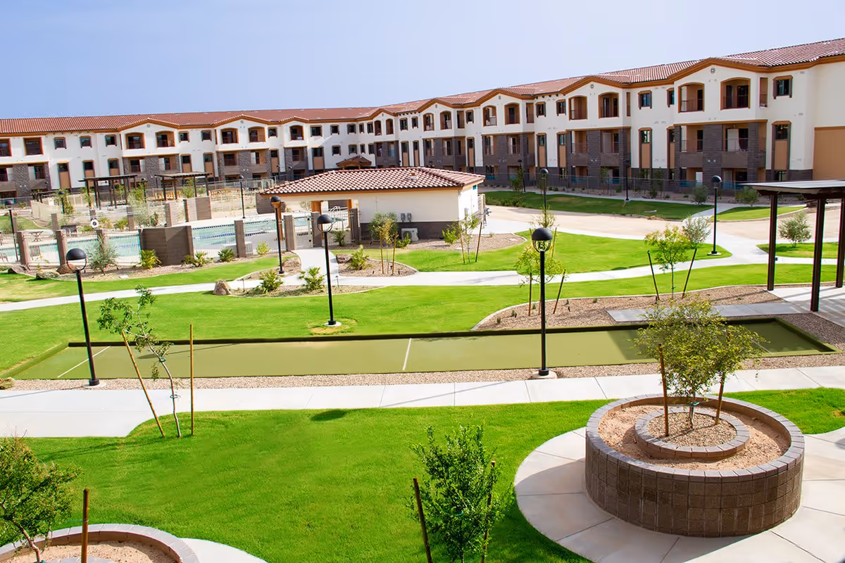 Outdoor view of Fellowship Square Surprise senior living facility showing a large, multi-story building with balconies, a well-maintained green lawn, walking paths, small trees, lamp posts, a bocce ball court, and a fenced swimming pool area.