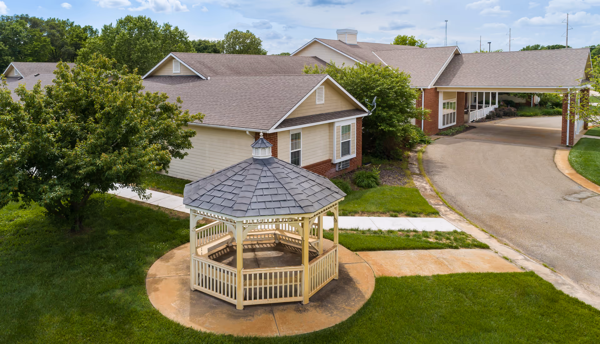 A white wooden gazebo on a circular concrete pad in the lawn in front of a single-story assisted living building with a covered driveway.