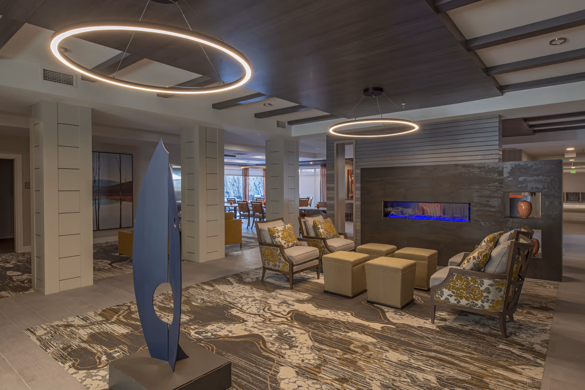 A modern senior living facility common area with comfortable armchairs featuring floral cushions, square ottomans, a decorative blue sculpture, and a contemporary fireplace with blue flames. The space has circular pendant lights hanging from a dark wood ceiling and a patterned carpet covering the floor. In the background, there is a dining area with tables and chairs near large windows.