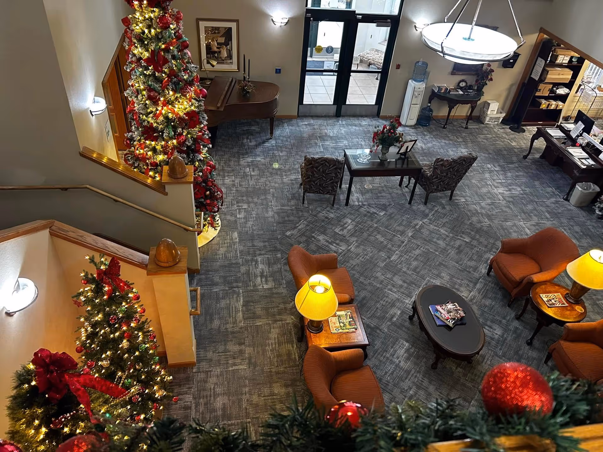 Interior view of a senior living facility lobby decorated for Christmas with two decorated Christmas trees, comfortable seating with armchairs and tables with lamps, a grand piano, and a reception desk near the entrance doors.