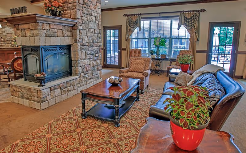 A cozy living room area with a stone fireplace, a wooden coffee table, a leather sofa, two patterned armchairs, and a large window with curtains. There are decorative plants on the tables and a patterned rug on the floor.