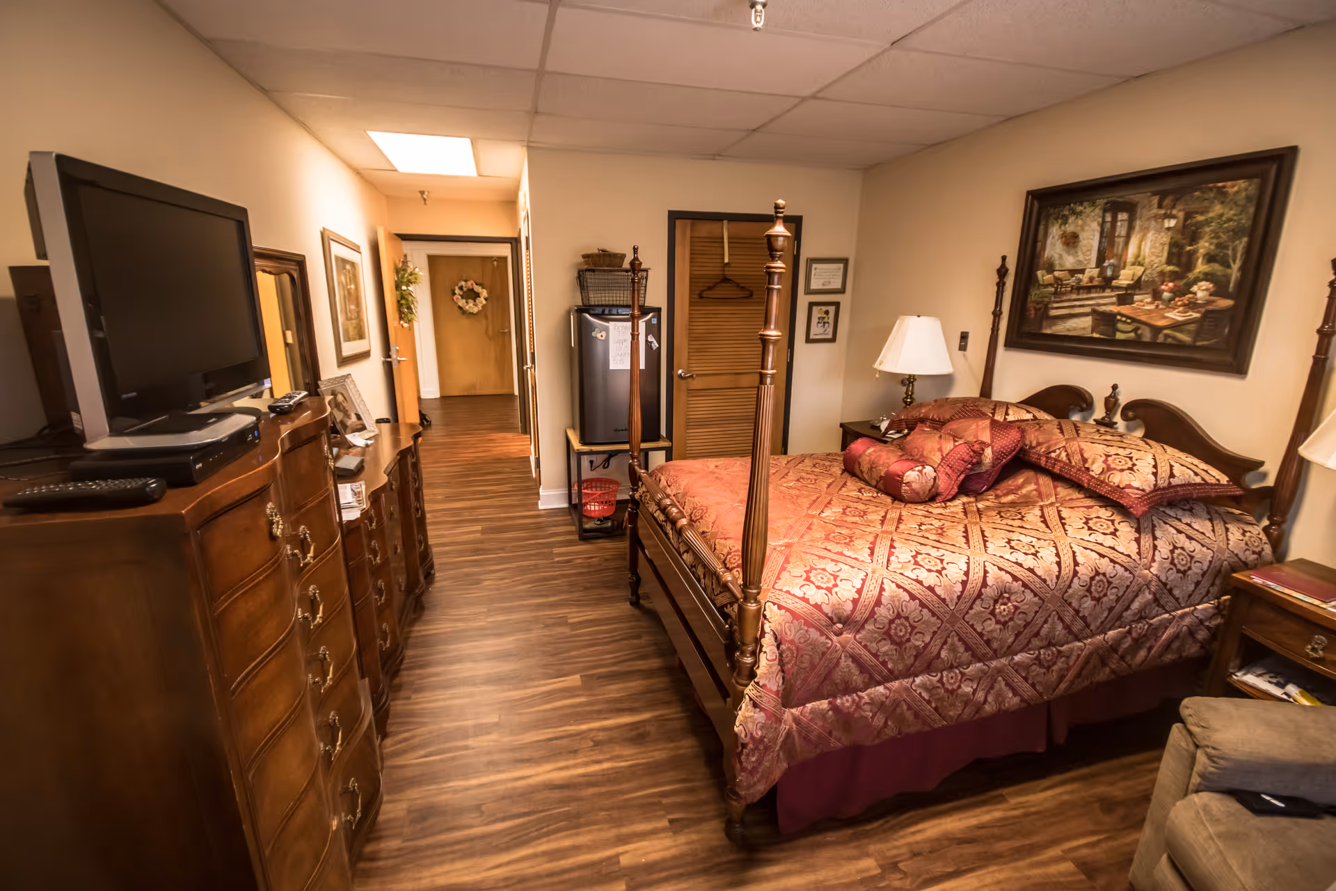 A cozy bedroom in a senior living facility with a wooden four-poster bed covered in a patterned red and gold bedspread and multiple pillows. There is a wooden dresser with a flat-screen TV on top, a nightstand with a lamp, and framed artwork on the walls. The room has wood flooring and a hallway leading to a door decorated with a wreath.