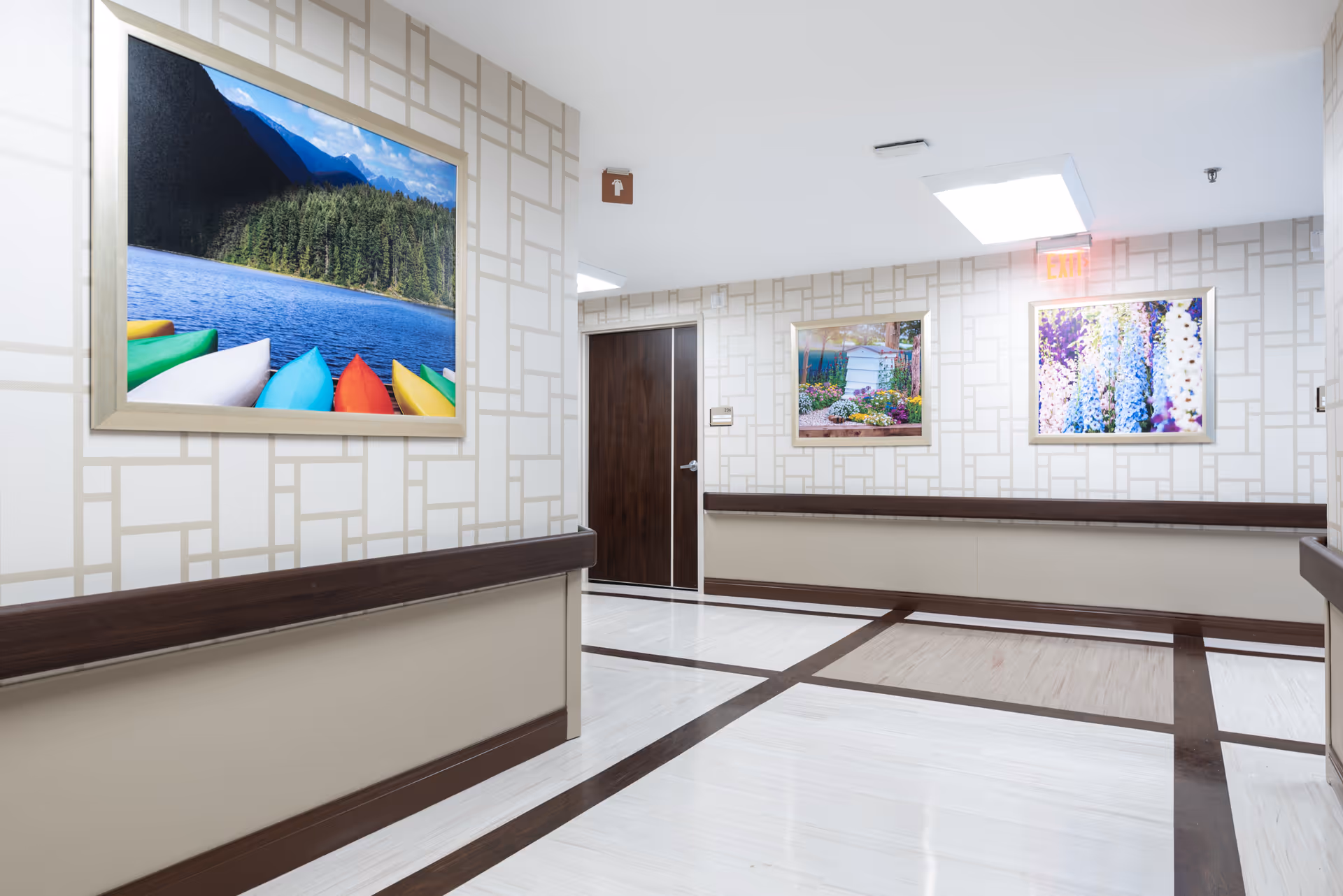A clean, well-lit hallway in a senior living facility with patterned wallpaper, dark wood trim, and three framed nature photographs on the walls. The floor has a light wood pattern with dark wood inlays, and there is a closed dark wood door at the end of the hallway.