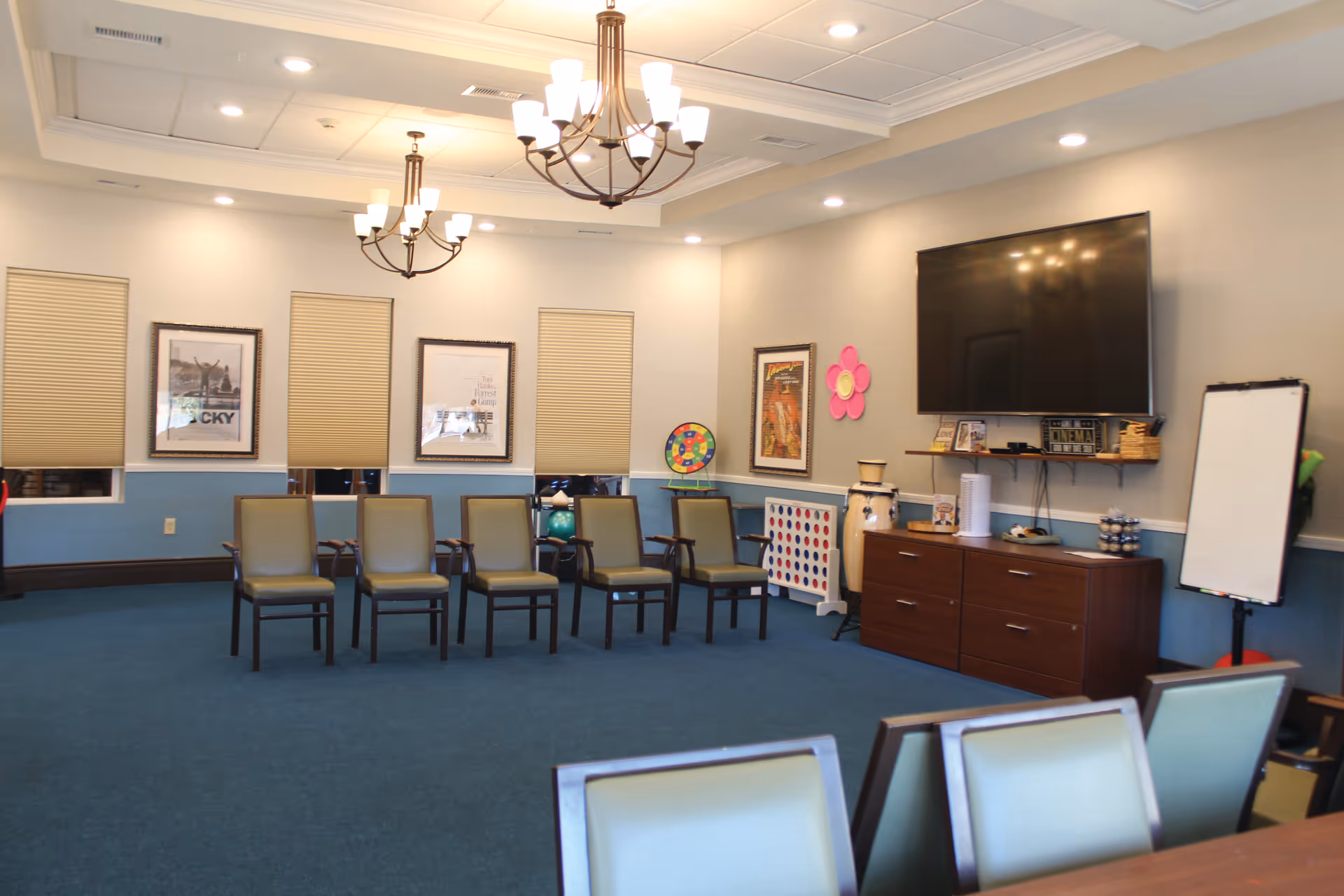 A memory care common room with multiple chairs, a wall TV, and a cabinet.