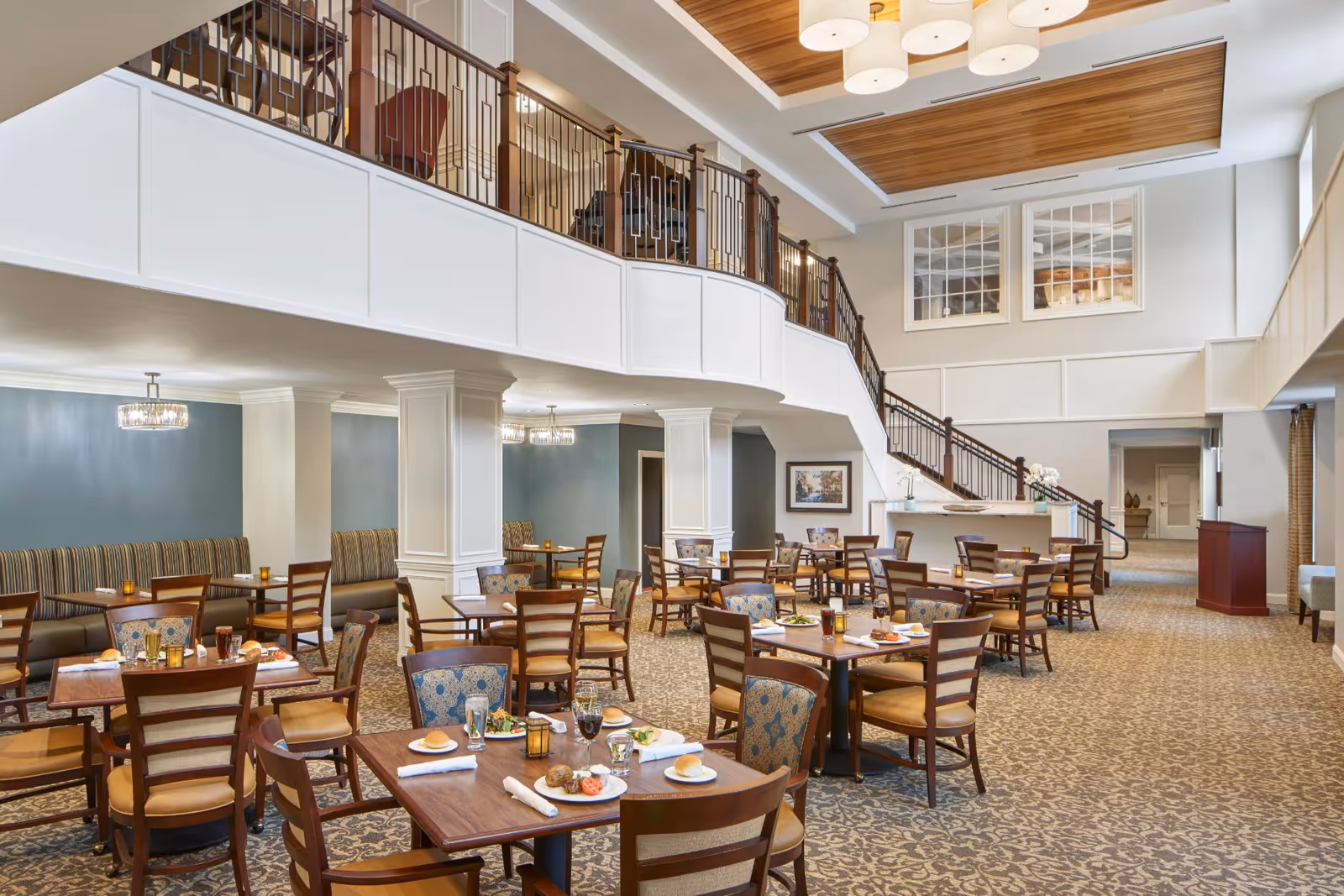 Spacious dining area with multiple set tables and chairs, a mezzanine balcony and staircase beneath high ceilings.