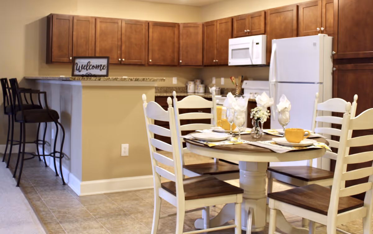 A cozy kitchen and dining area featuring wooden cabinets, a white refrigerator, microwave, and stove. The dining table is set with four white chairs, place settings including plates, glasses with napkins, and yellow cups. There are two bar stools at the kitchen counter, and a small decorative sign that says 'Welcome' is placed on the counter.