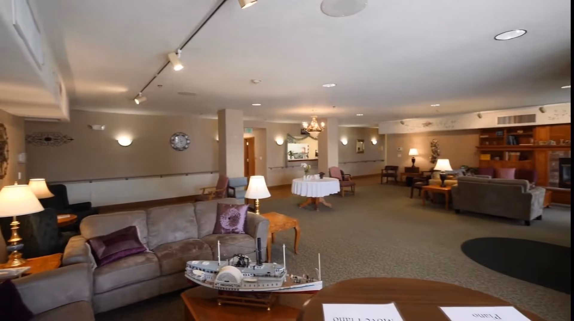 A spacious and well-lit living room area in a senior living facility with beige walls and carpeted floor. The room features multiple seating arrangements including sofas with purple cushions, armchairs, and wooden side tables with lamps. A model ship is displayed on a wooden table in the foreground. The back wall has a clock, wall sconces, and a built-in wooden bookshelf with decorative items. There is a round table covered with a white tablecloth in the center of the room, and a chandelier hanging from the ceiling.