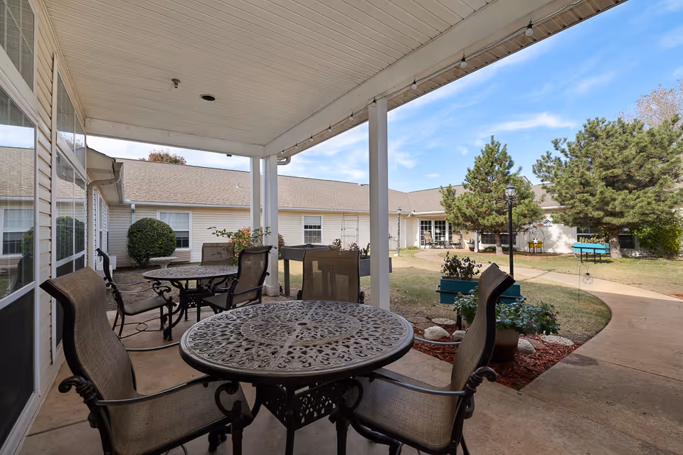 Covered outdoor patio area with metal tables and chairs, overlooking a courtyard with trees, plants, and a paved walkway. The building surrounding the courtyard has beige siding and multiple windows.